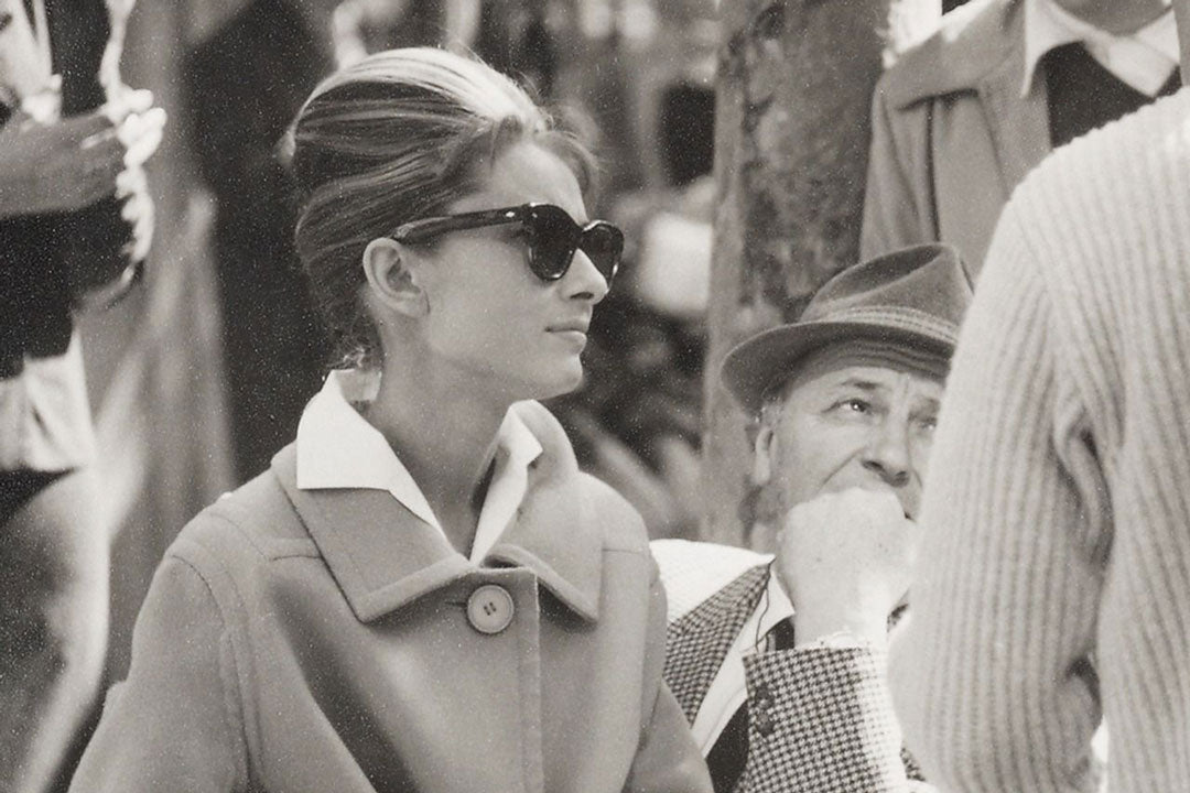 Side view of Audrey Hepburn sitting down wearing beige coat and large sunglasses