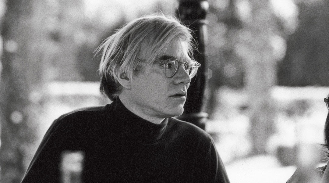 Side view of artist Andy Warhol wearing black polo neck and round clear glasses frame