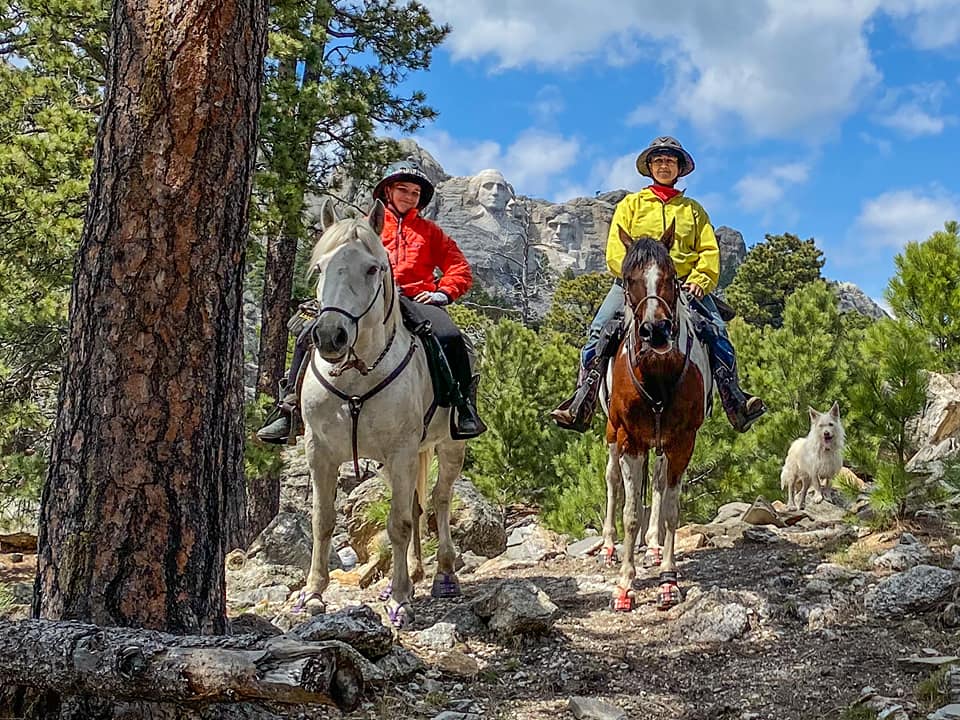 A woman and her daughter riding horses wearing Scoot Boots in the forrest in front of Mt. Rushmore