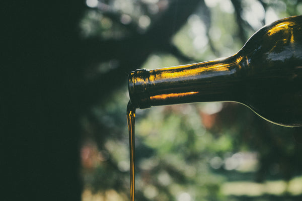 The sun shines through a dark green bottle as an reddish amber oil is poured out, there are trees in the background.