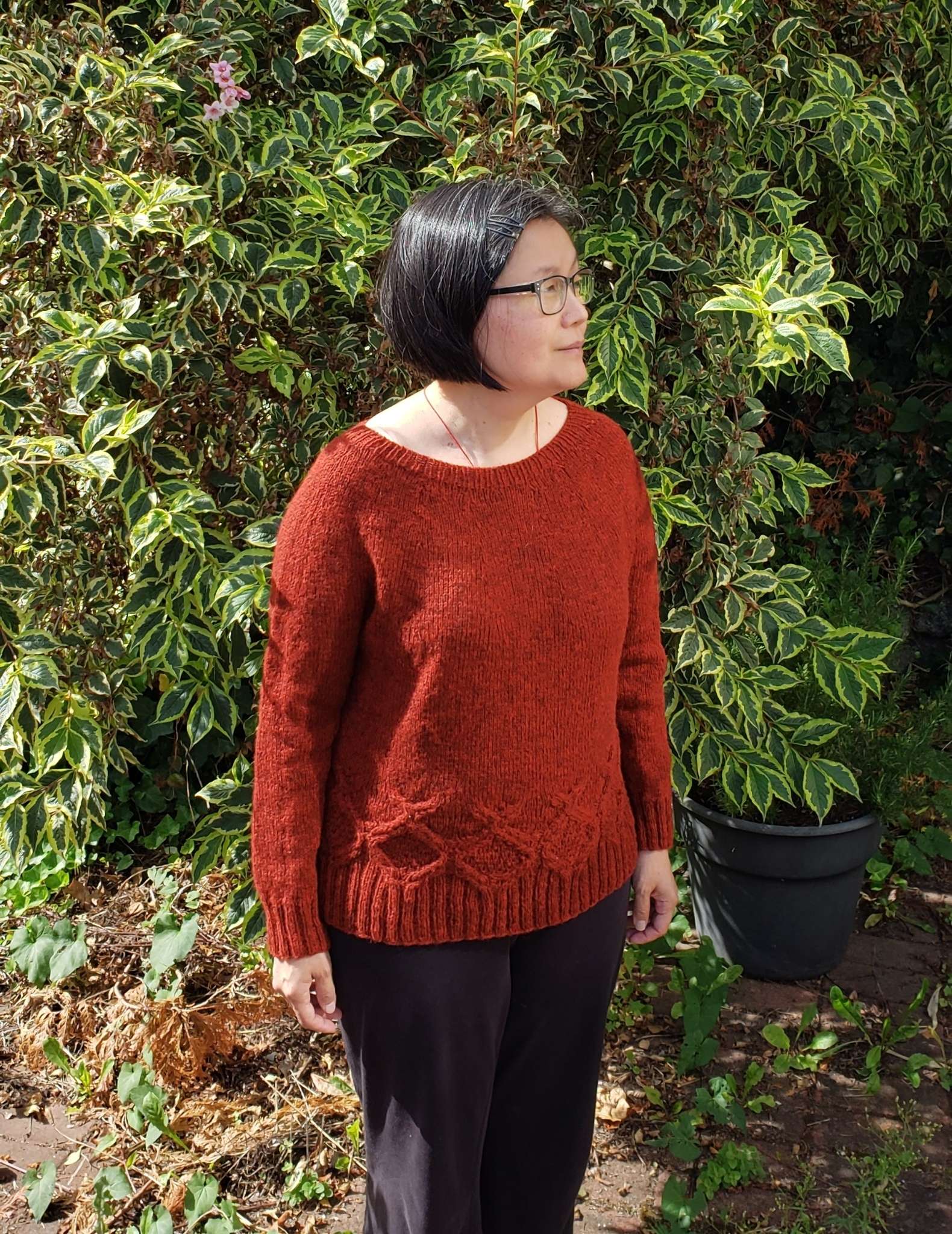 a model with dark hair wearing an orange cabled sweater stands in front of a bush, looking to the side