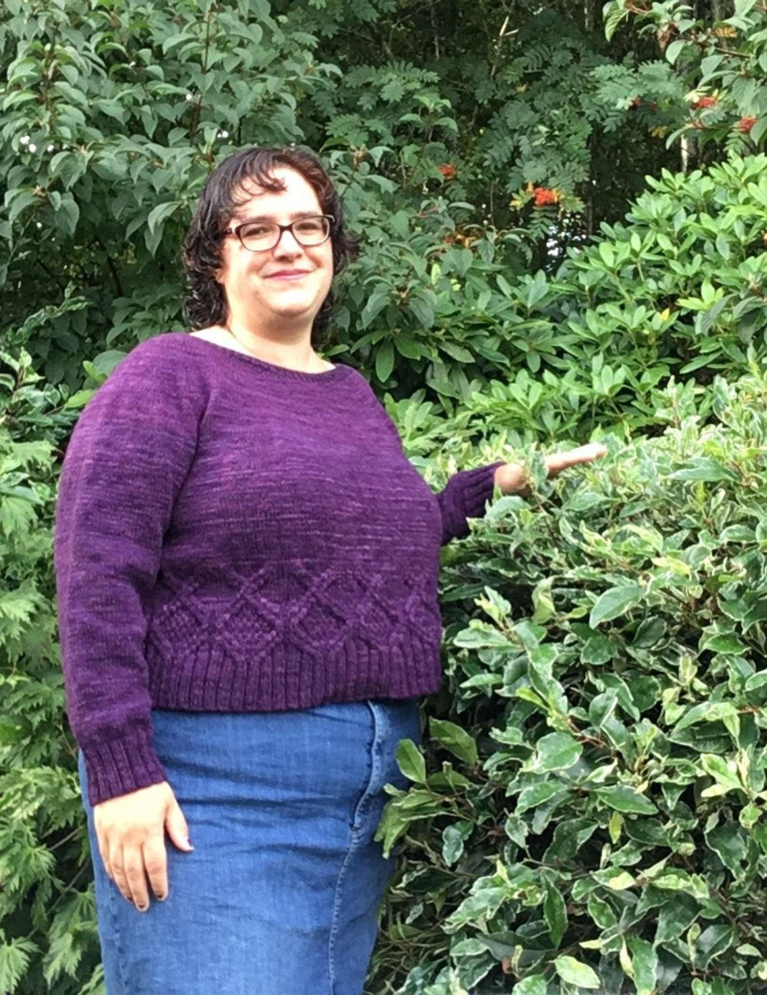 a model with dark hair wearing a purple sweater, standing in front of a bush with one arm raised