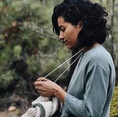 Image of a light brown skinned woman knitting with her working yarn wrapped over her neck