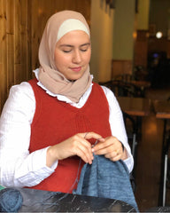 Image of an Arabic woman wearing a hijab and a red vest knitting a blue project