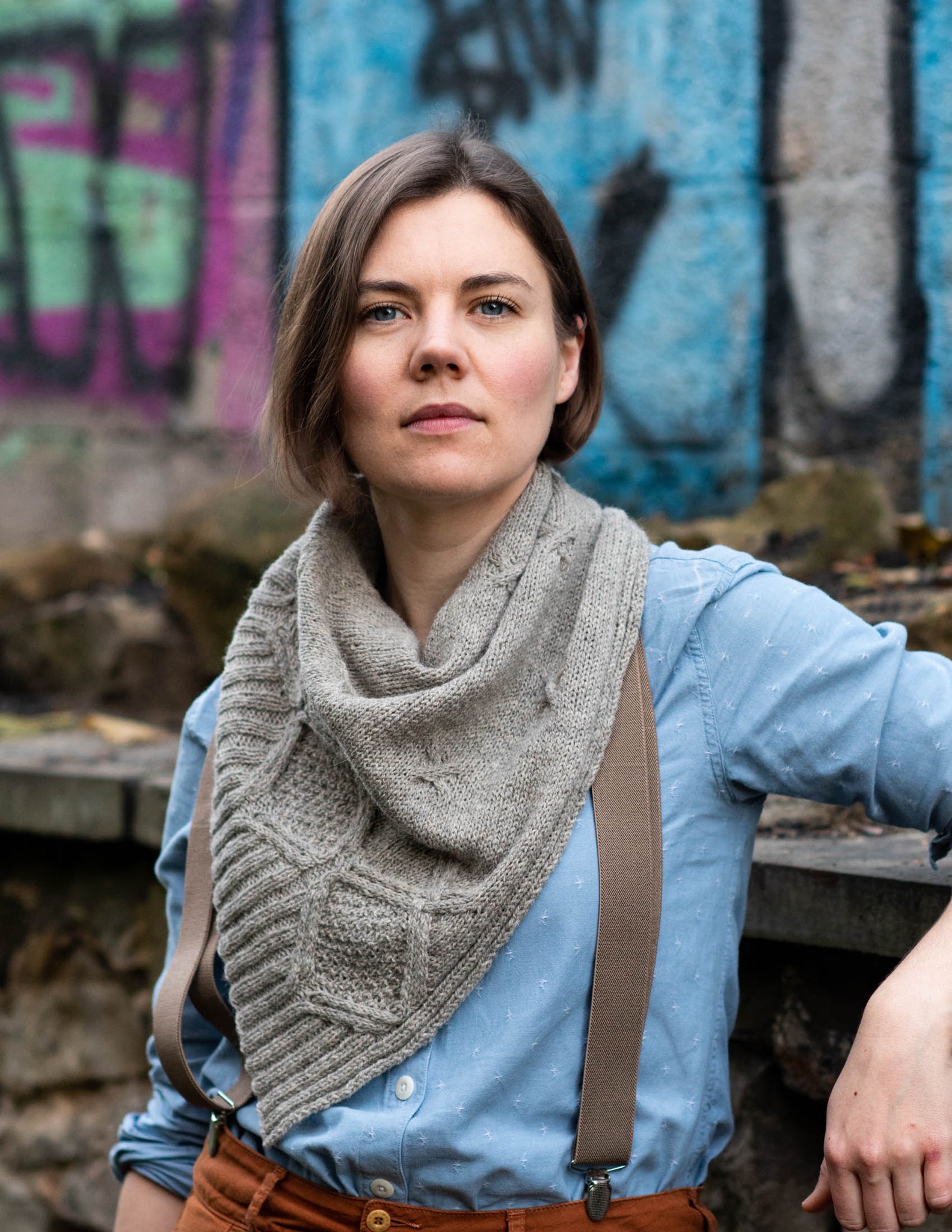 A white woman wears a grey cabled shawl wrapped round her neck, a blue shirt with braces and brown trousers. She leans one one elbow in front of a graffitied background.  
