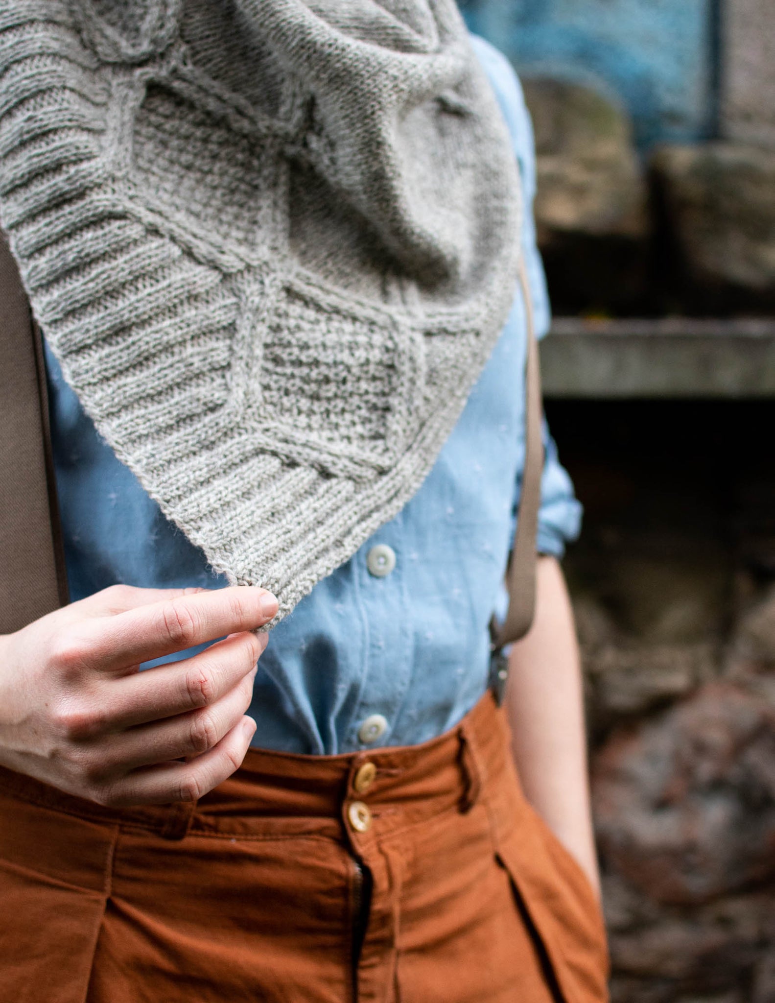 a close of of a cabled edge of a grey shawl, the corner of which is held out by a white hand. The model wears a blue shirt with braces and brown trousers.