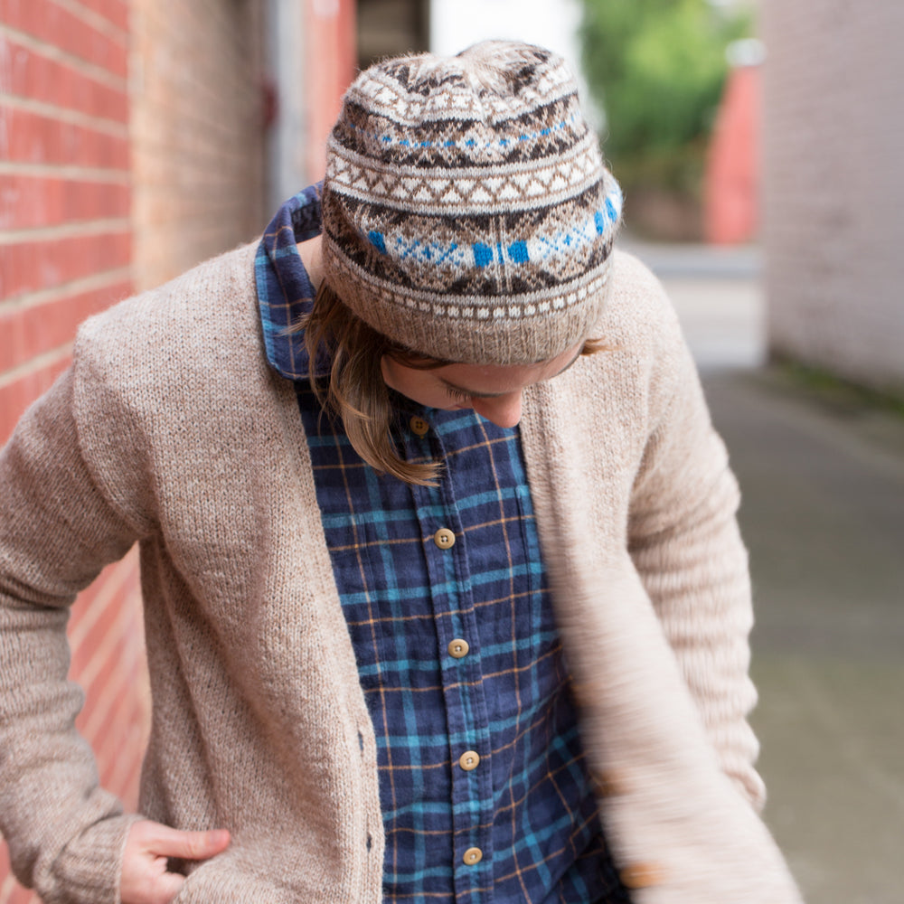 Ysolda, a white woman with chin-length brown hair, stands next to a brick wall looking at the ground. She is wearing a brown and grey Fair Isle hat with a blue accent detail, a blue plaid shirt and brown cardigan