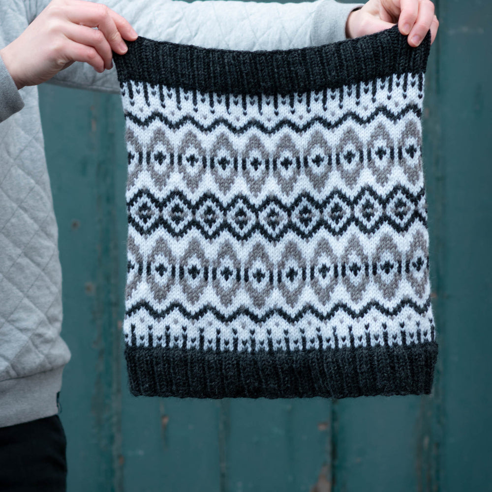 Two white hands hold a black, white, and grey cowl outstretched. the cowl is stranded colourwork with strong oval and diamond motifs