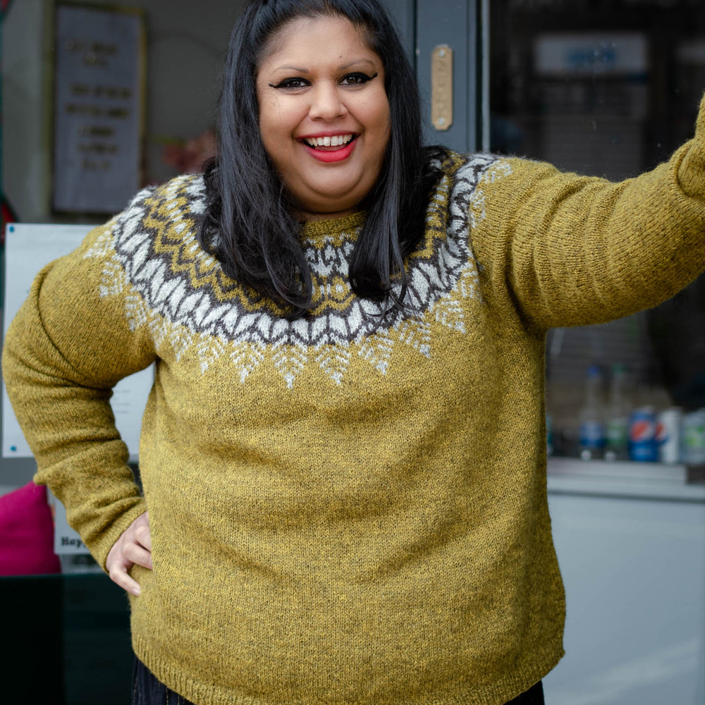 A south asian woman with mid length dark hair stands in a doorway looking towards the right of the frame. She is wearing  an earthy green knit sweater with a dark brown and light grey colourwork yoke.