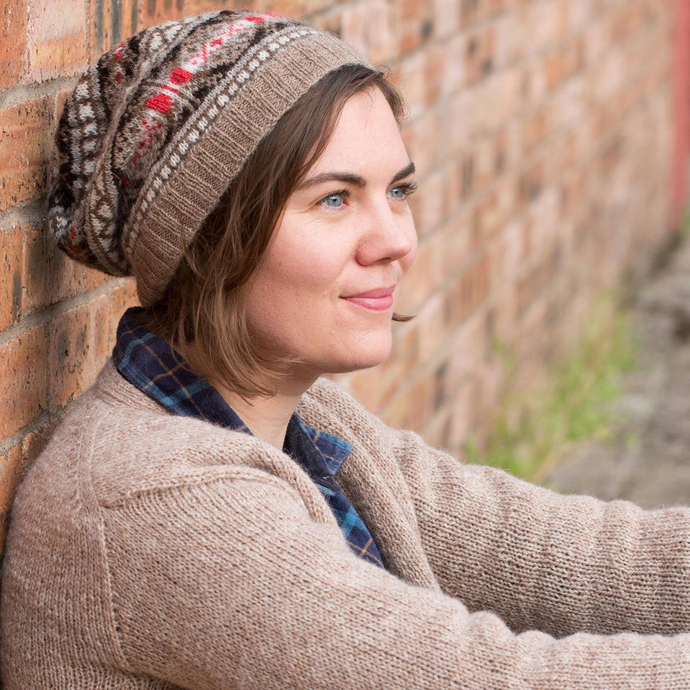Ysolda, a white woman with chin-length brown hair, sits against a brick wall looking to the side. she is wearing a brown jumper and a brown and red fair isle hat. 