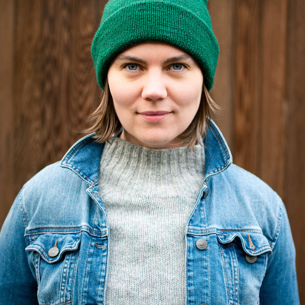 Ysolda, a white woman, stands looking directly at the camera with a small smile. She is wearing a bright green watchcap, grey jumper, and denim jacket
