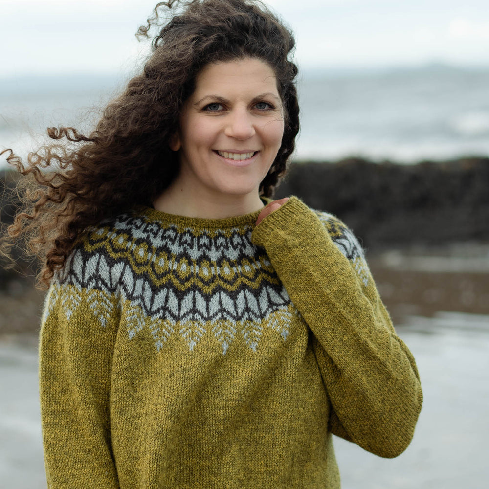 A white woman with long curly brunette hair stands on the beach smiling at the camera with the sea behind her. She is wearing an earthy green knit sweater with a dark brown and light grey colourwork yoke. Her left arm is folded up and her hand is near her neck holding the cuff of her sweater.