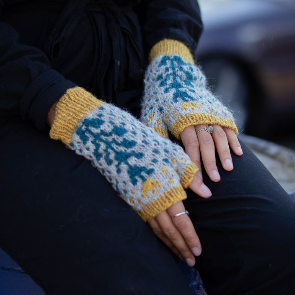 Close up of someone in black with their hands in their lap. They are wearing  bold grey background and blue geometric botanic wool fingerless mitts with yellow trim at the cuff, and fingers.. 