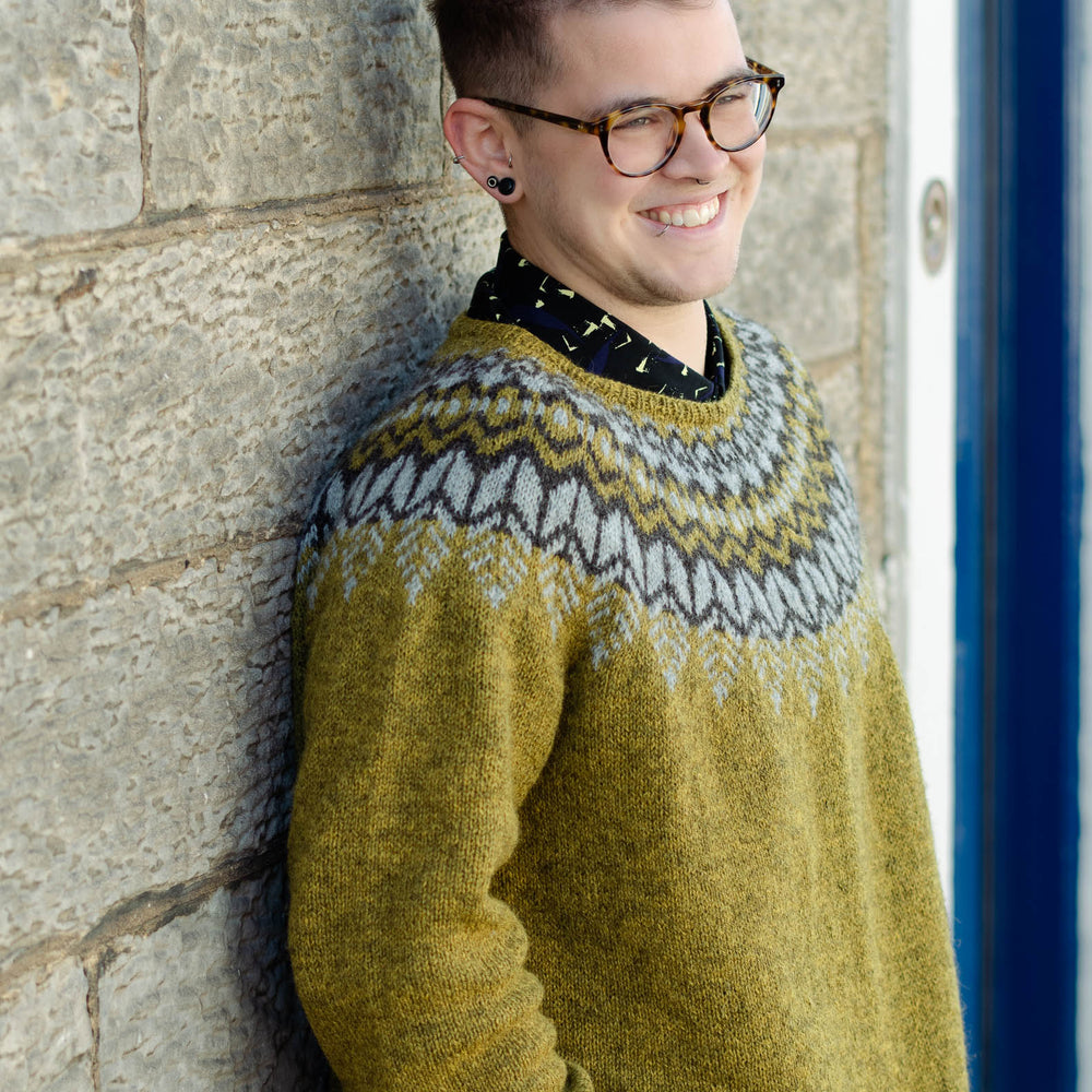 A white man leans casually against a light coloured stone wall, he has short hair, is wearing dark glasses and smiles at the camera. He is wearing an earthy green knit sweater with a dark brown and light grey colourwork yoke.