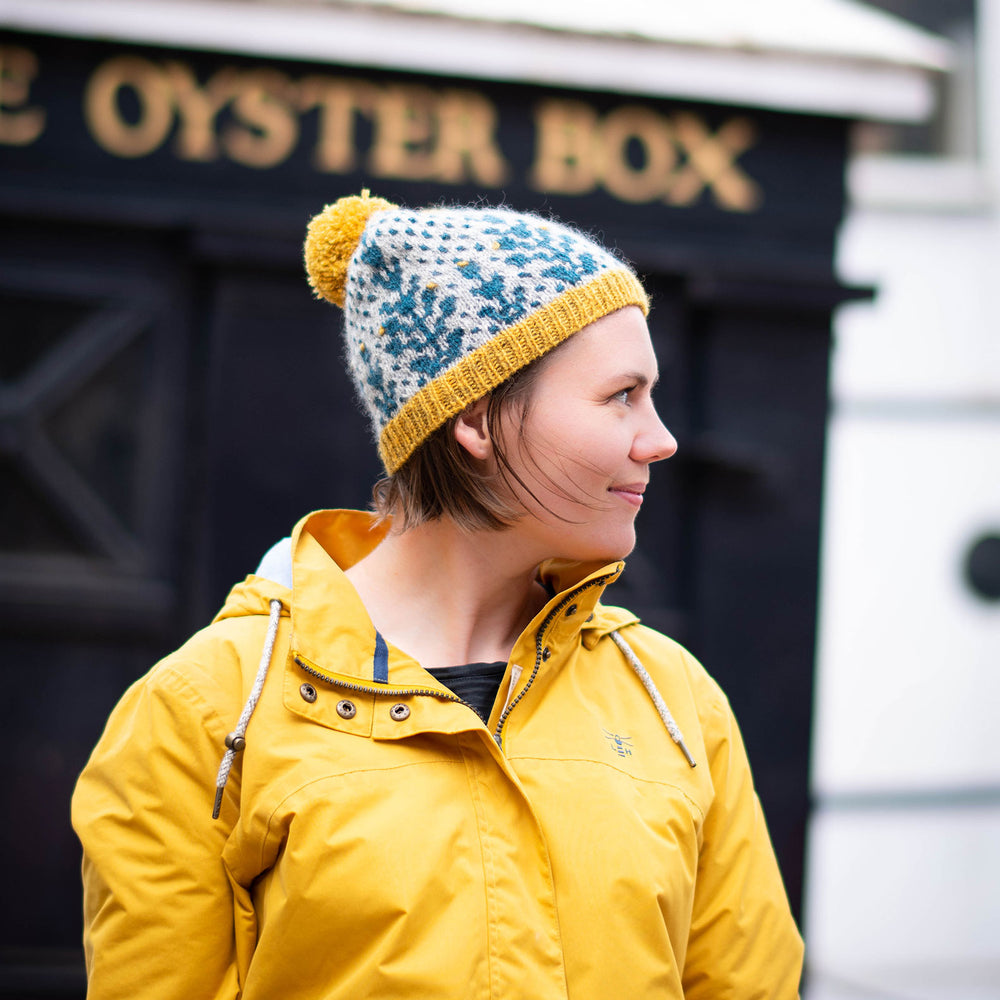 A white woman stands facing  to the right of the frame in front of a black painted restaurant front with gold letters &#039;Oyster Bar&#039;.. The are wearing a yellow hooded jacket with the hood down and wearing a bold grey background and blue geometric botanic wool hat with yellow trim and pompom. 