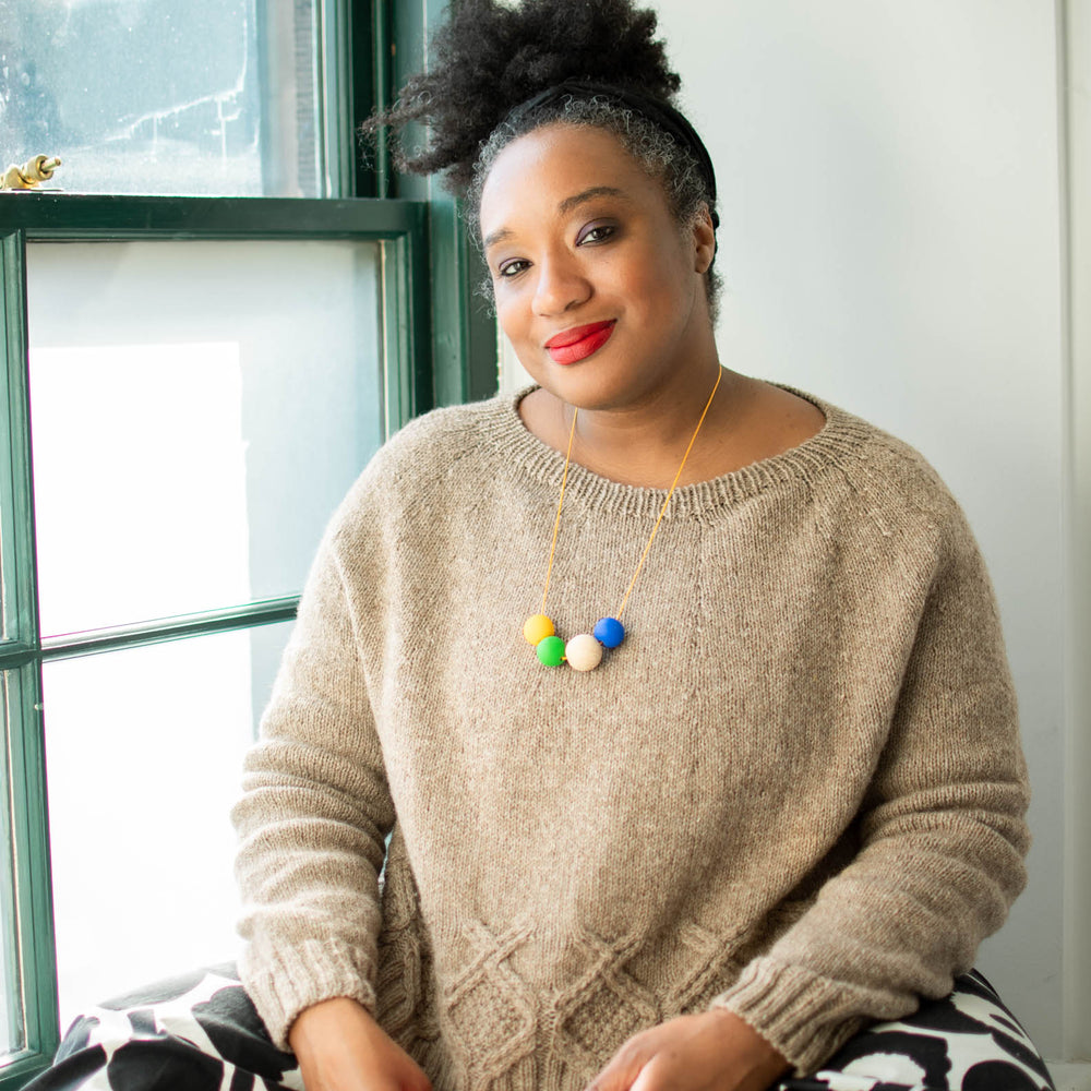 A black woman with their hair pulled up sits crossed legged on a window sill. She&#039;s wearing a cream coloured wool sweater with a round yoke and cables around the body just above the hem.