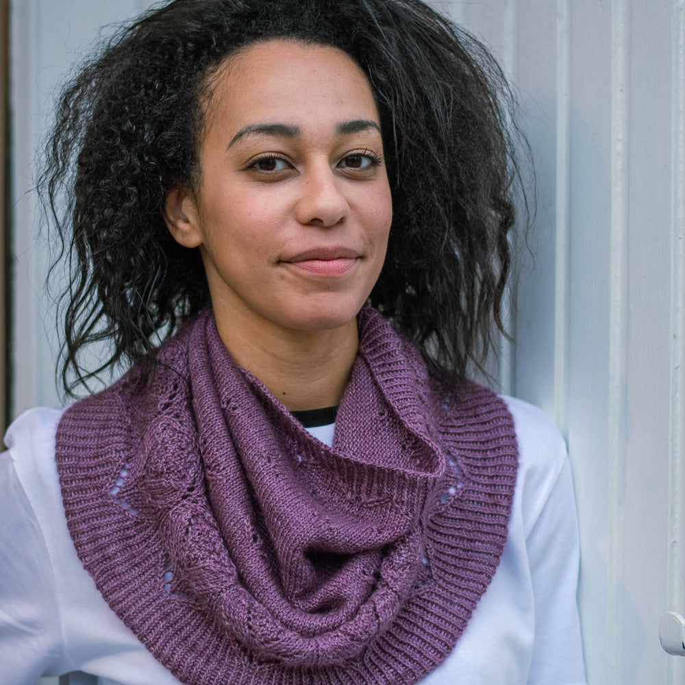 A black woman leans casually against a white wooden wall. She&#039;s wearing a deep purple knit cowl and a white t-shirt.