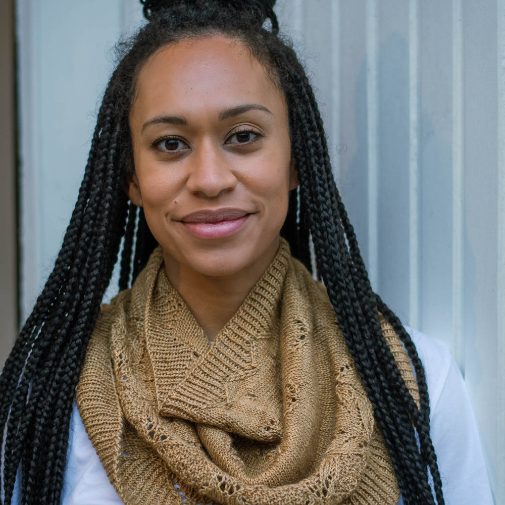 A black woman with long braids, leans casually against a white wooden wall. She&#039;s wearing a caramel coloured knit cowl and a white t-shirt.