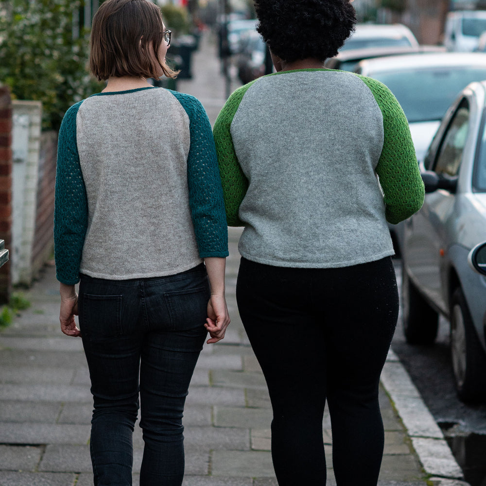 Two women stand with their back to the camera on a city sidewalk back lit by the sunset. Both are wearing black jeans and baseball style kit sweaters. The white woman on the left has a chin length bob, the sleeves of her top are teal. The black woman on the right has natural hair and the sleeves of her top are green.