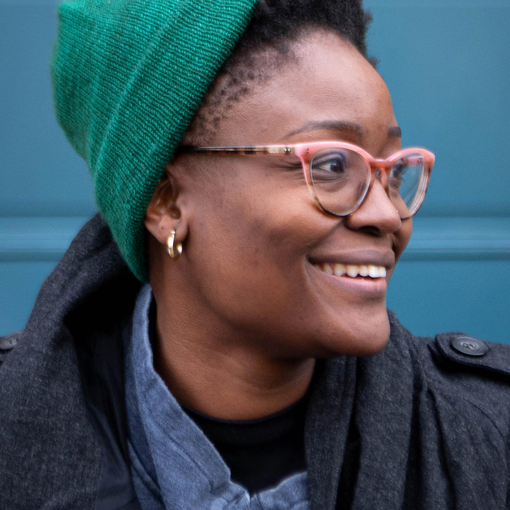 A Black woman looks to the side smiling. She is wearing a green beanie hat with natural hair peeking out the front. She is wearing brown and pink glasses, a denim shirt, and dark grey coat