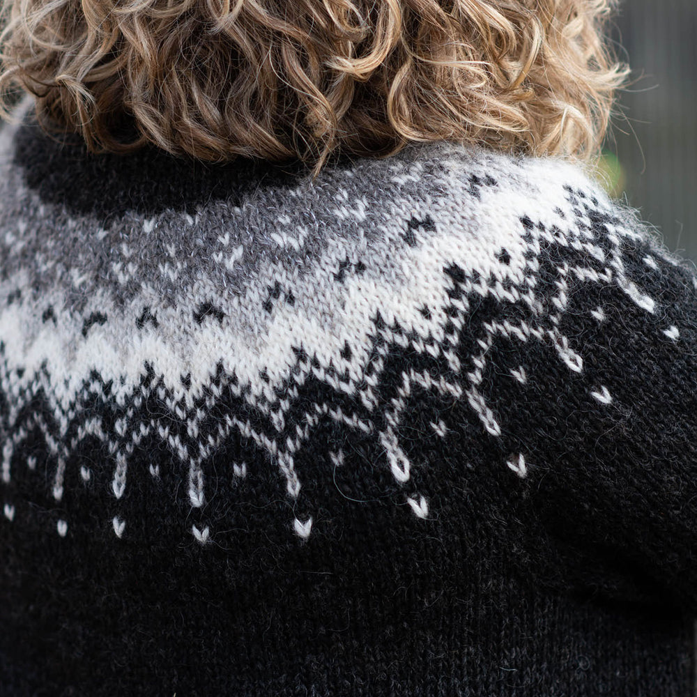 Cropped image of the back yoke of the Bleideag pattern, black sweater with a grey, silver, and white colourwork yoke. The person wearing the sweater has curly shoulder length blond hair.