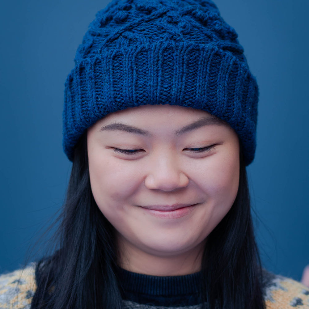 Portrait image of a young asian woman wearing a blue cable beanie with a folded brim. She has long dark hair, that hangs down over the front a icelandic yoked sweater. The back ground of the image is blue.