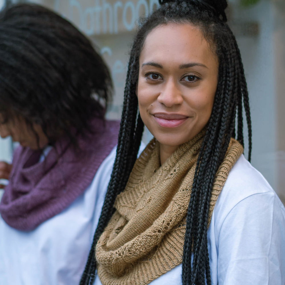 Two black women stand casually in front of a store window. The woman in the foreground has long braids, is wearing a caramel coloured knit cowl over a white t-shirt. The woman in the background has natural hair and she&#039;s wearing a deep purple knit cowl and a white t-shirt.
