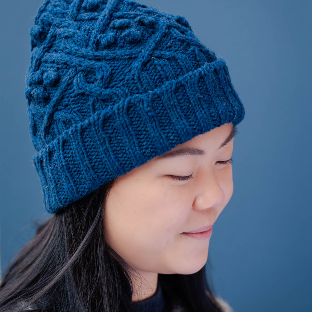 Profile image of a young asian woman wearing a blue cable beanie with a folded brim. She has long dark hair, that hangs down over the front an icelandic yoked sweater. 