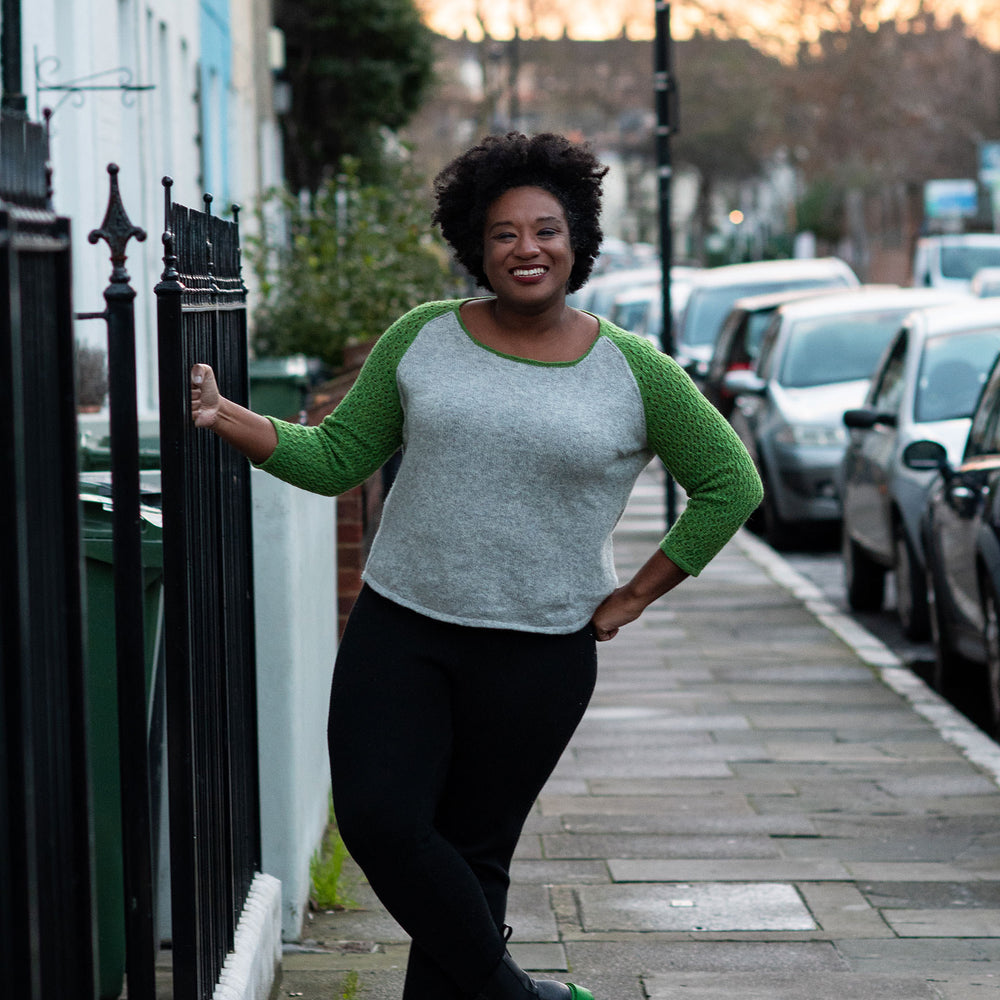 A black woman stands hanging on to the railing on a city sidewalk back lit by the sunset. They are wearing black jeans and a baseball style knit sweater with a grey body and green sleeves. They have natural hair and big smile.