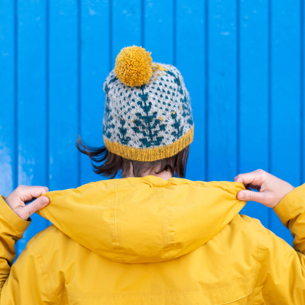 A person stands facing away from the camera towards a wooden slated wall painted blue. The are wearing a yellow hooded jacket with the hood down. They are holding the edges of the hood. They are wearing a bold grey background and blue geometric botanic wool hat with yellow trim and pompom. 