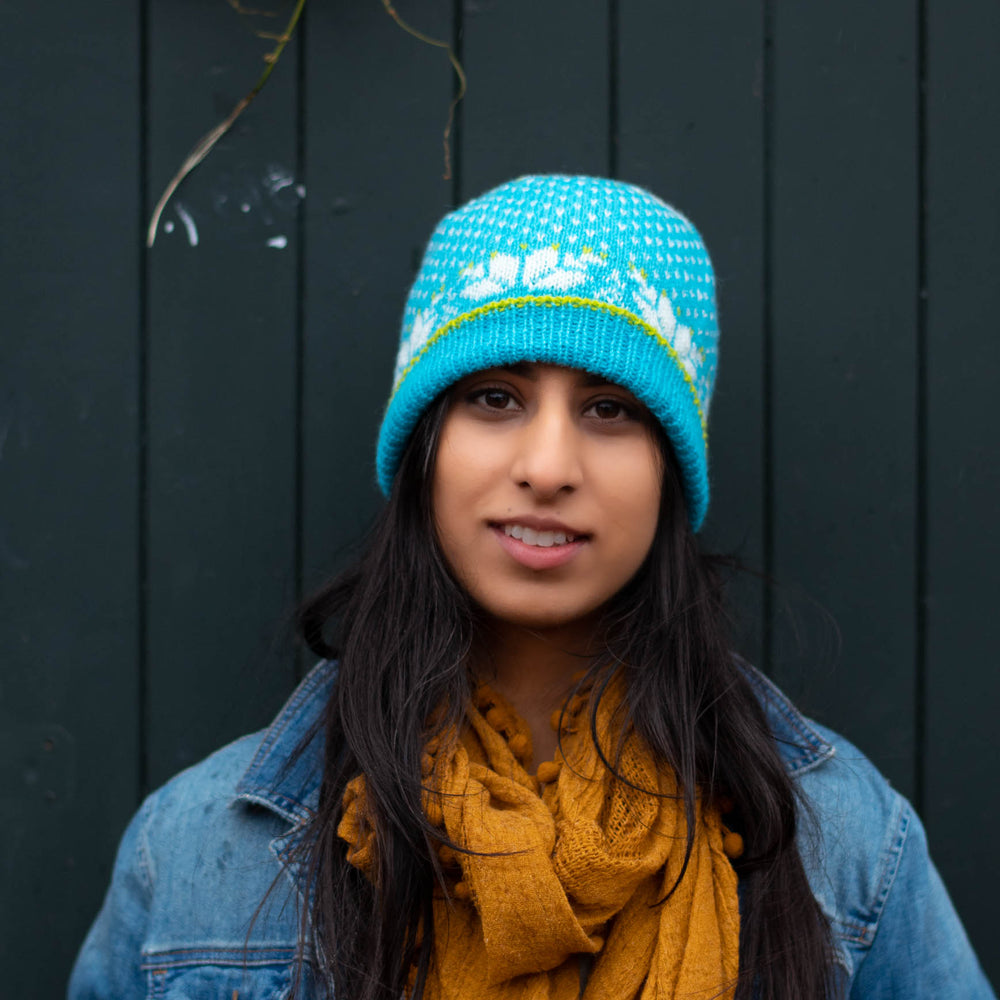 South Asian woman faces the camera smiling, she wears a bright blue stranded colourwork hat with bold star motifs, jean jacket, and yellow scarf.