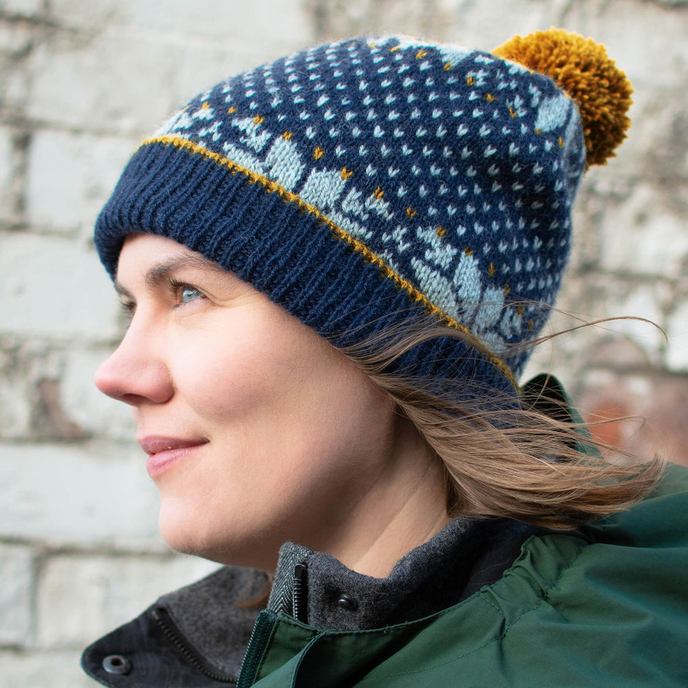 White woman with short brown hair stands in profile, wearing a dark blue stranded colourwork hat with a yellow pom pom, and she has on a green jacket