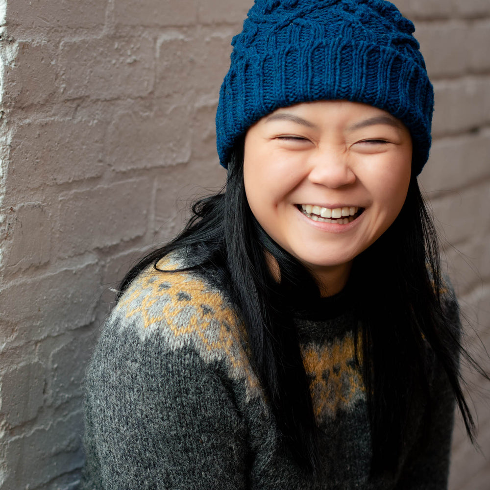 Portrait image of a young asian woman wearing a blue cable beanie with a folded brim. She has long dark hair, that hangs down over the front an icelandic yoked sweater. She&#039;s in front a white brick wall, she exudes joy.