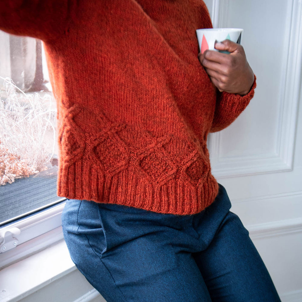 A person leans against a window sill. Wearing an orange wool sweater with cables above the ribbing, with dark blue trousers.