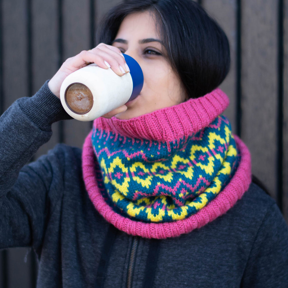 South Asian woman drinks from a reusable mug, she is wearing a stranded colourwork cowl in vibrant colours - neon yellow, neon pink, and deep blue