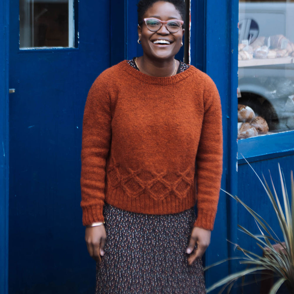 A black woman with short hair and glasses, stands in front of a doorway painted blue. She&#039;s laughing and facing the camera, wearing an orange wool sweater with a round yoke and cabled detail around the waist over a dark coloured mini floral print maxi dress.
