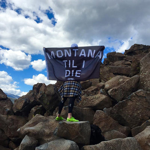 A black flag with white lettering that says Montana Til I Die. Held by a young woman in work out clothing standing on a hillside.