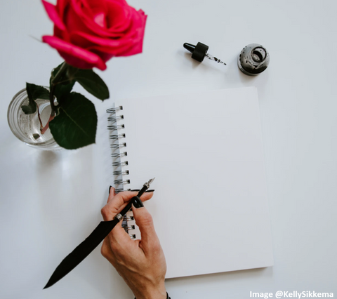 a hand holding an old fashioned ink quill and resting on a white ringed notepad with a red rose on a white table