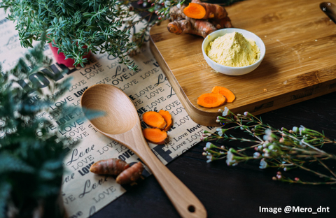 wooden spoon resting on a table with fresh herbs and turmeric slices around it and a wooden cutting board