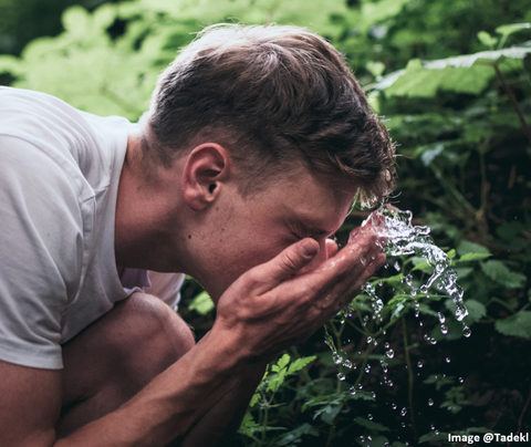 young blonde man washing his face with water and green leaves of a forest behind him