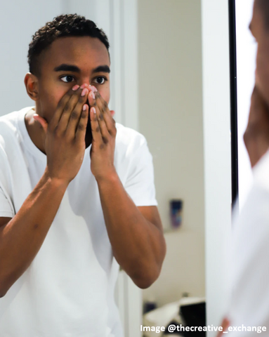 man looking in mirror touching his face with both hands, covering his nose and mouth as if putting on face lotion