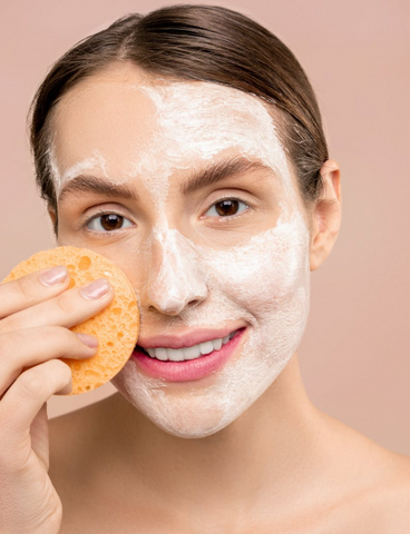 woman smiling with hair pulled back and a sponge in her hand with a white cream mask on her face
