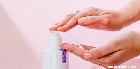 two neatly trimmed manicured hands pressing on a lotion bottle pump and white lotion squirting out in an arc shape