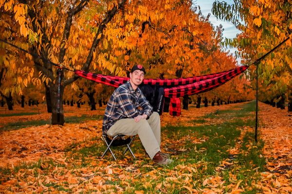 man sitting in a grove of trees in the fall, in front of a plaid hammock