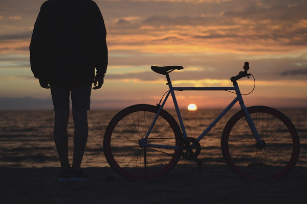 person standing next to a bicycle on the beach staring at the sun setting over the water