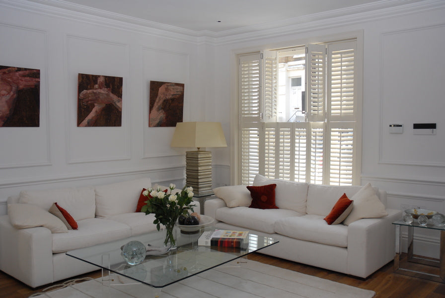 modern living room with glass table,  two white square armchairs, modern pictures on the white wall, cream open curtains and full height white window shutters