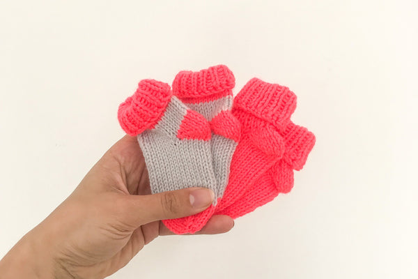 Women’s hand holding two pairs of baby socks in neon coral and grey