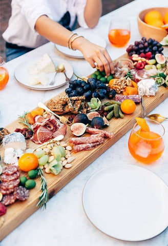 Cured meats, crackers, cheese, olives, pickles, fresh berries & figs, dried fruit and nuts on a wooden board with orange cocktail on the side