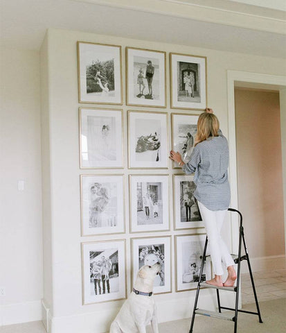 Woman hanging framed photos in wall to ceiling grid gallery style.