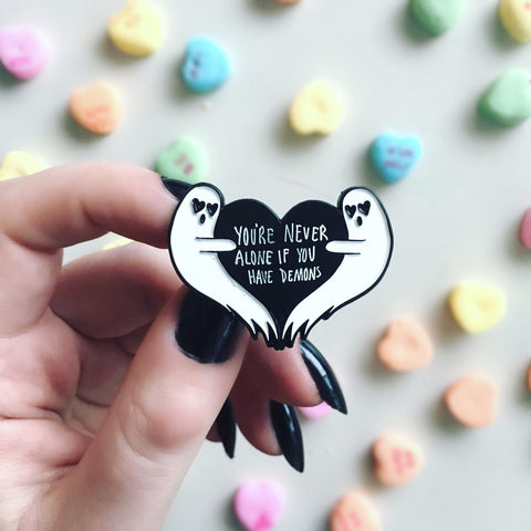 A woman holding an enamel pin of a black heart held by two ghosts. It has the funny quote, "You're Never Alone If You Have Demons". 